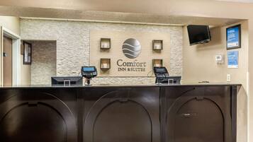 A reception desk at Comfort Inn & Suites with a stone wall backdrop, two ECHOCHIP payment terminals, a television mounted on the wall, and a framed informational sign.