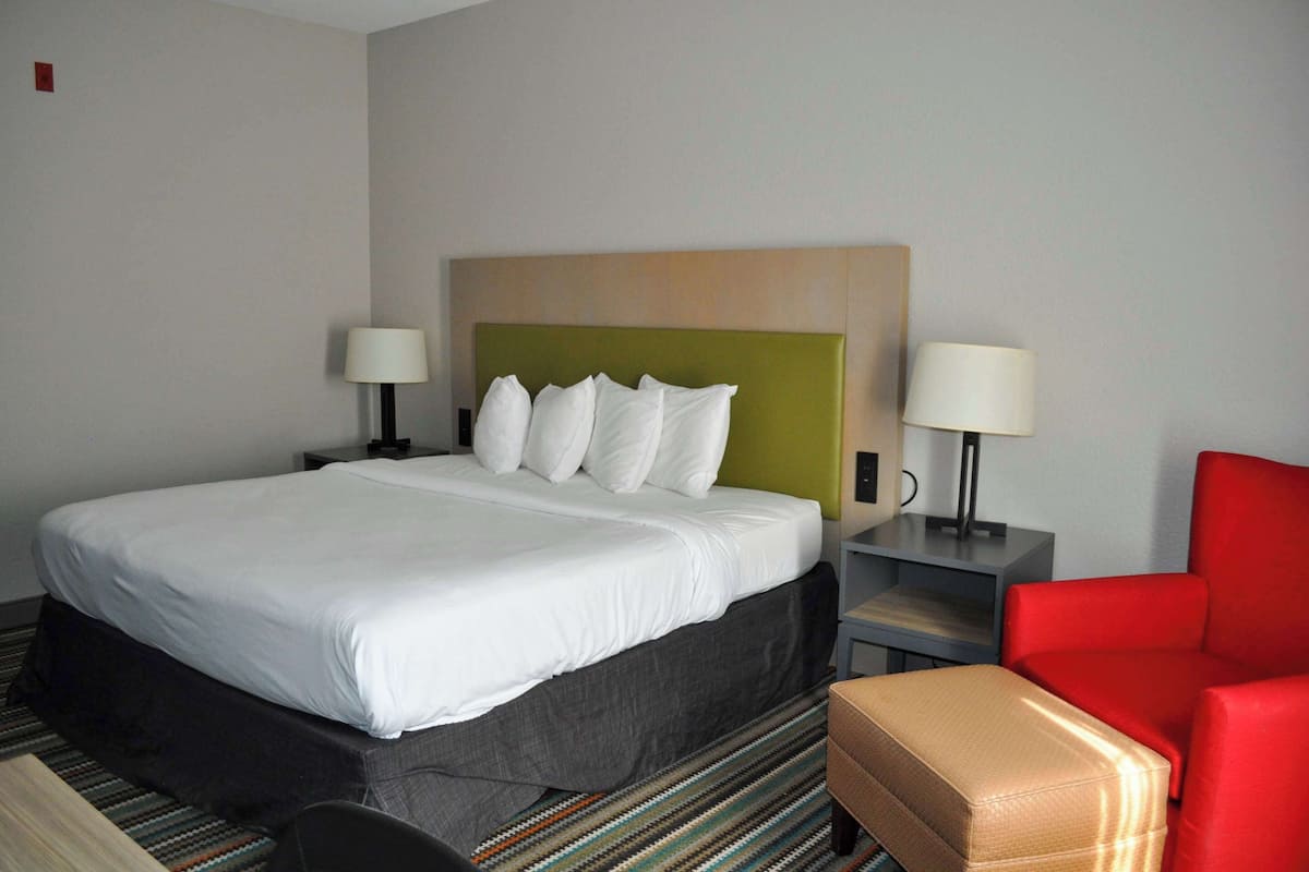 A hotel room with a large bed, two bedside tables, a red chair, and a small table with a lamp.
