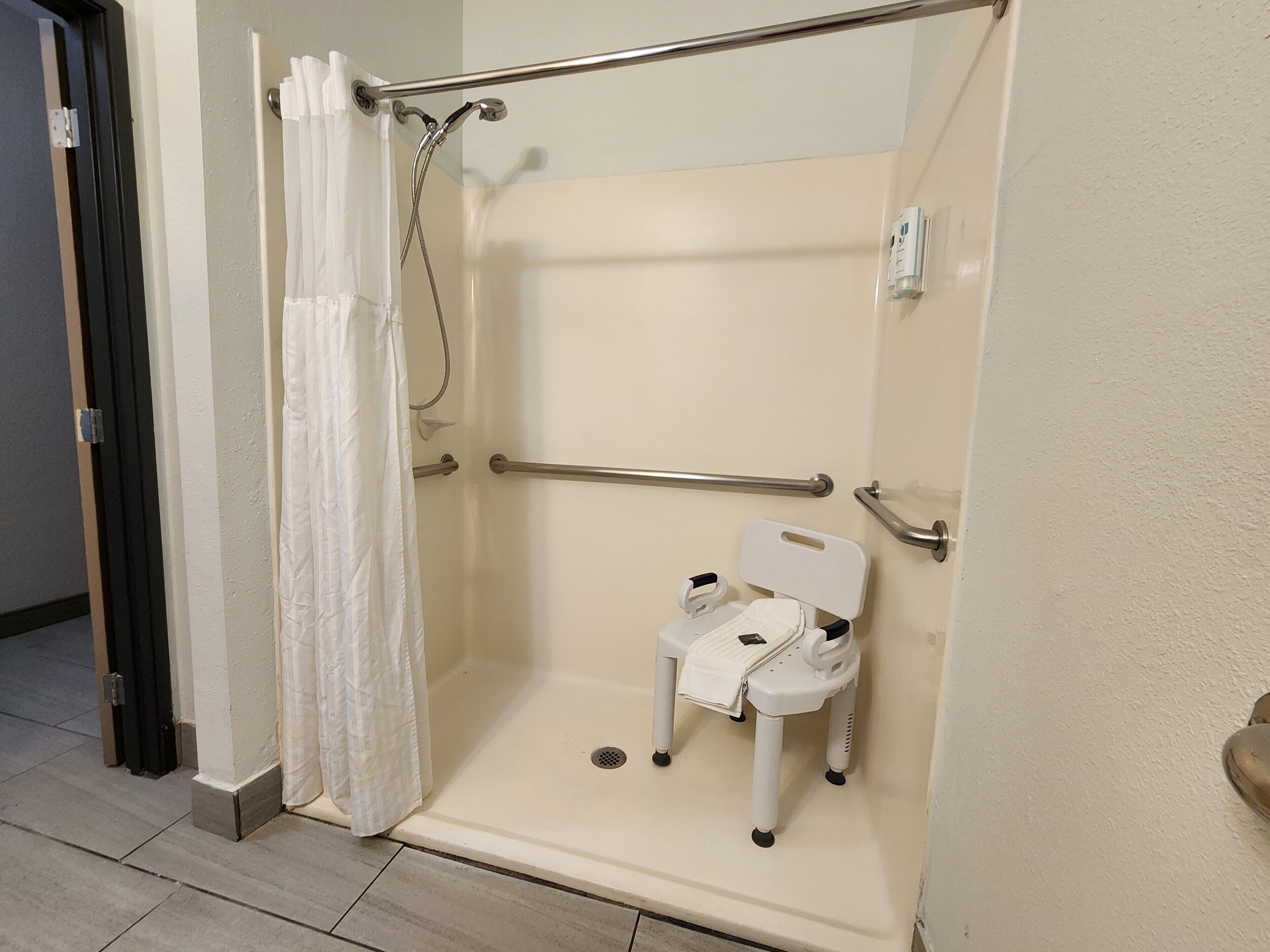 A wheelchair-accessible shower with a grab bar, a white shower chair, and a handheld showerhead.