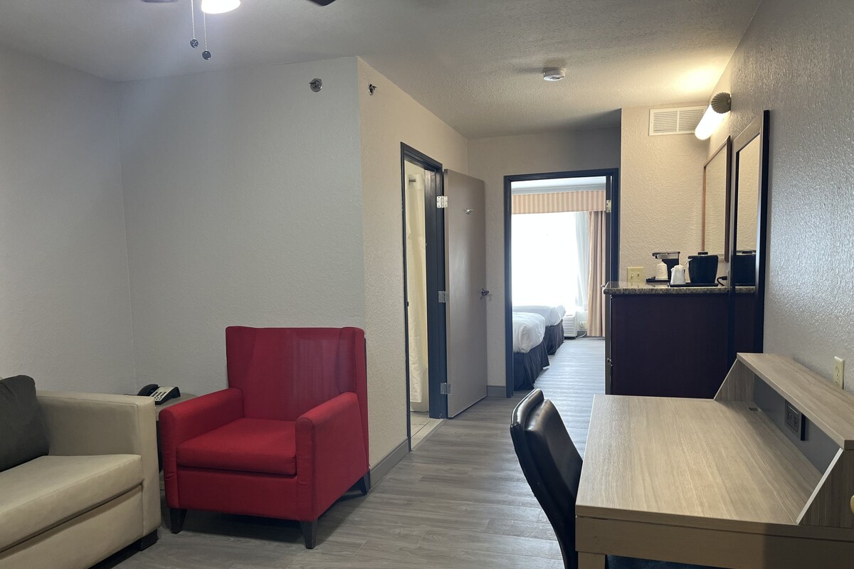 Ein Hotelzimmer mit einem roten Sessel, einem beigen Sofa, einem Holztisch und einer kleinen Küchenzeile.