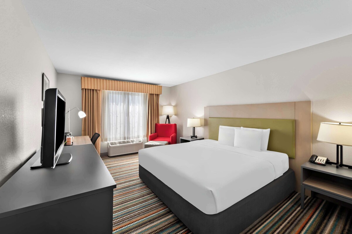 Ein Hotelzimmer mit einem großen Bett, einem Schreibtisch mit Flachbildfernseher, einem roten Sessel, einem Nachttisch mit Lampe und einem Fenster mit Vorhängen.