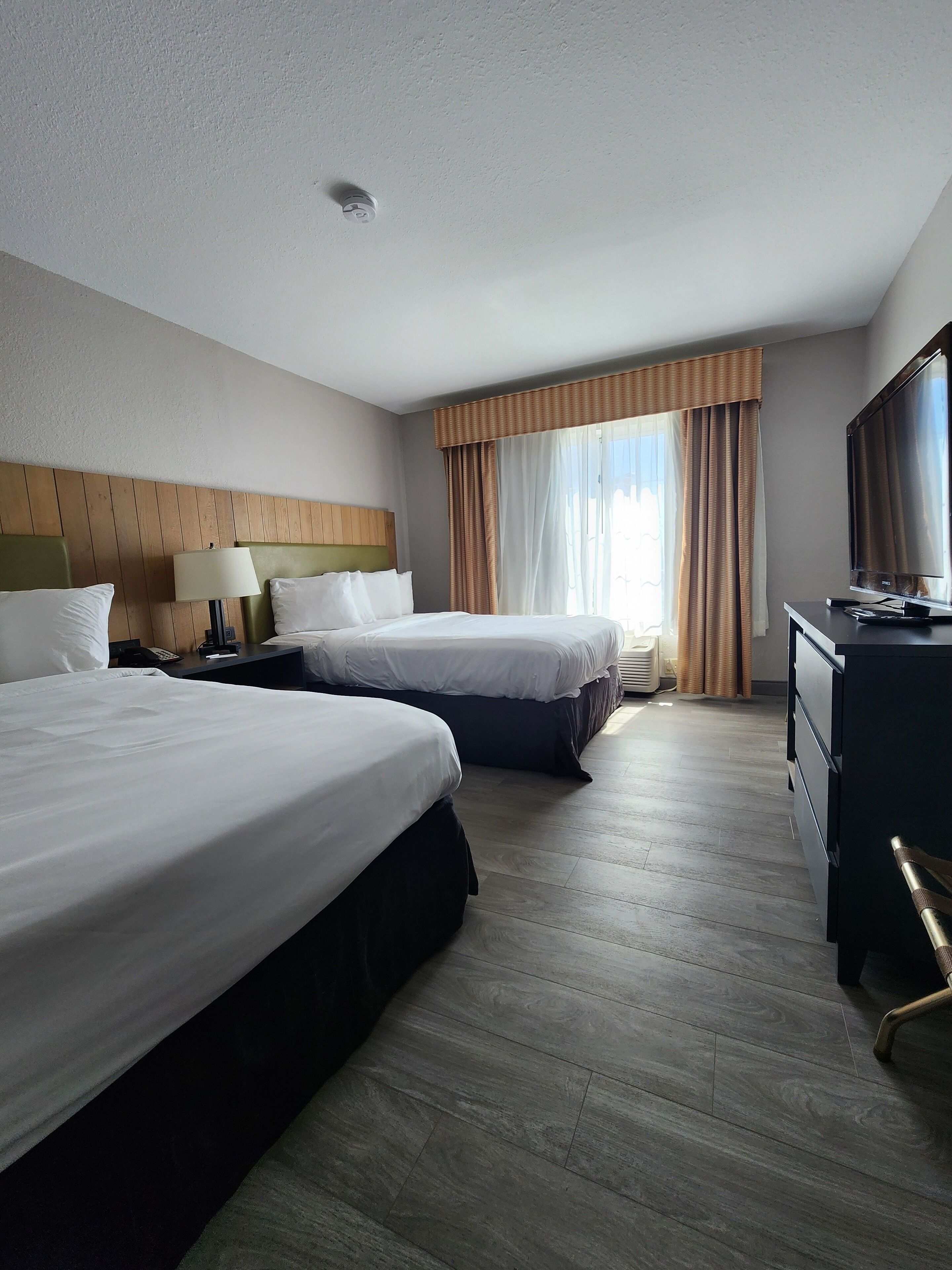 A hotel room with two beds, a desk with a lamp, a TV, and a large window with curtains.