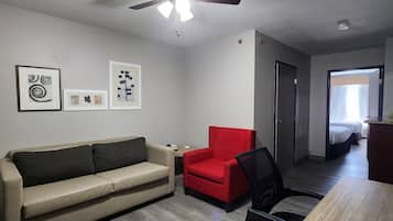 A room with a ceiling fan, a wooden table, a red chair, a beige sofa, and a bed in the background.
