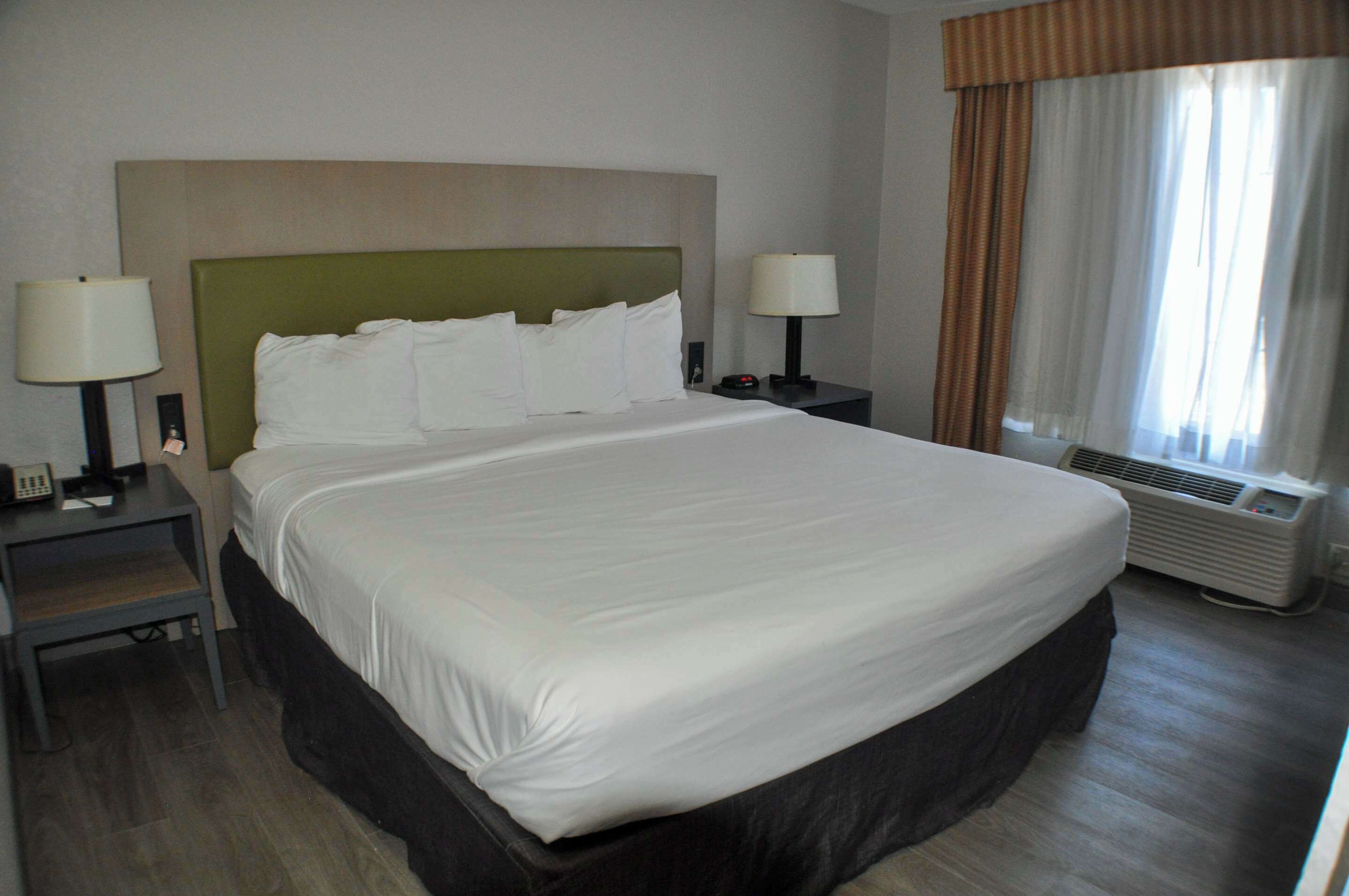 A hotel room with a large bed, two bedside tables with lamps, a window with curtains, and an air conditioning unit.