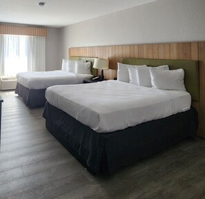 A hotel room with two beds, a wooden headboard, a nightstand with a lamp, and a window with curtains.