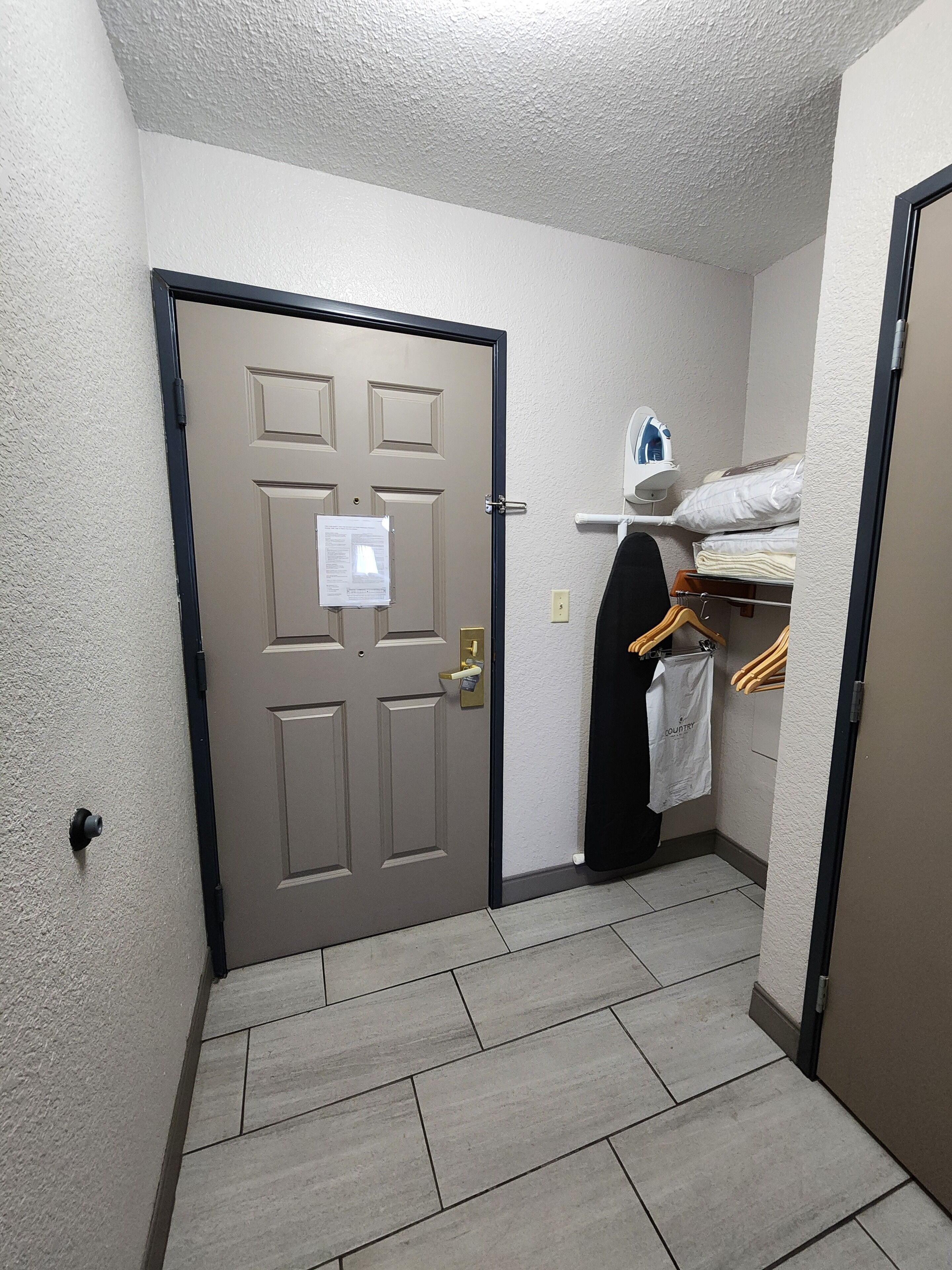 A narrow hallway with a beige door, a small ironing station with an iron and ironing board, and a rack with hangers and a white bag.