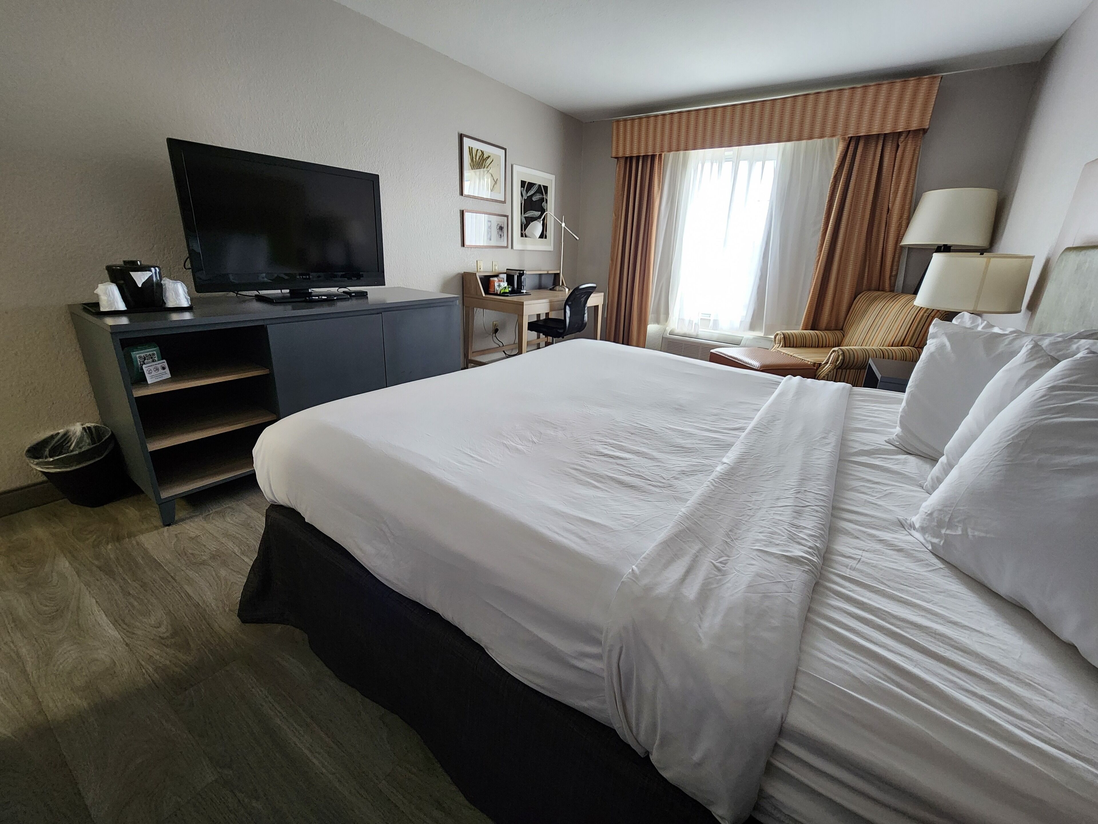 A hotel room with a large bed, a desk with a television, a chair, and a lamp.