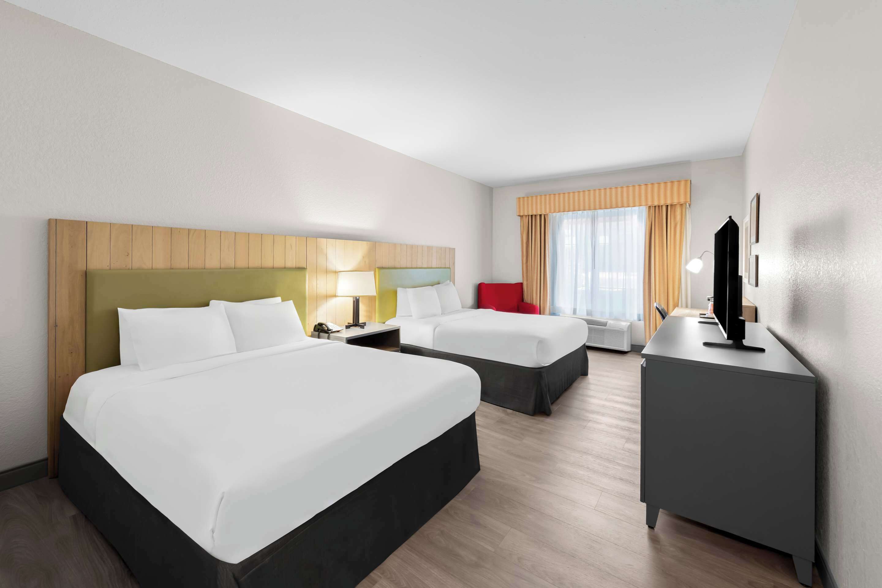 Quarto de hotel com duas camas, uma escrivaninha, uma cadeira, televisão e janela com cortinas.
