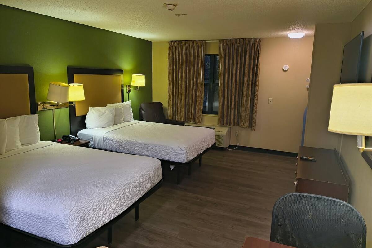 A hotel room with two beds, a desk, a chair, a lamp, and a window with curtains.