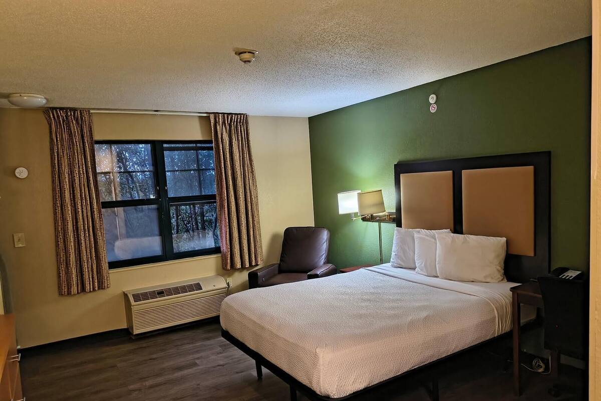 A hotel room with a bed, a chair, a desk, a lamp, and a window with curtains.