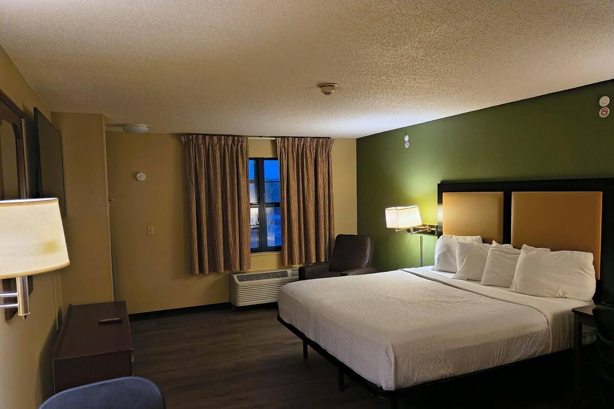A hotel room with a bed, a desk, a chair, a window with curtains, and a wall-mounted lamp.