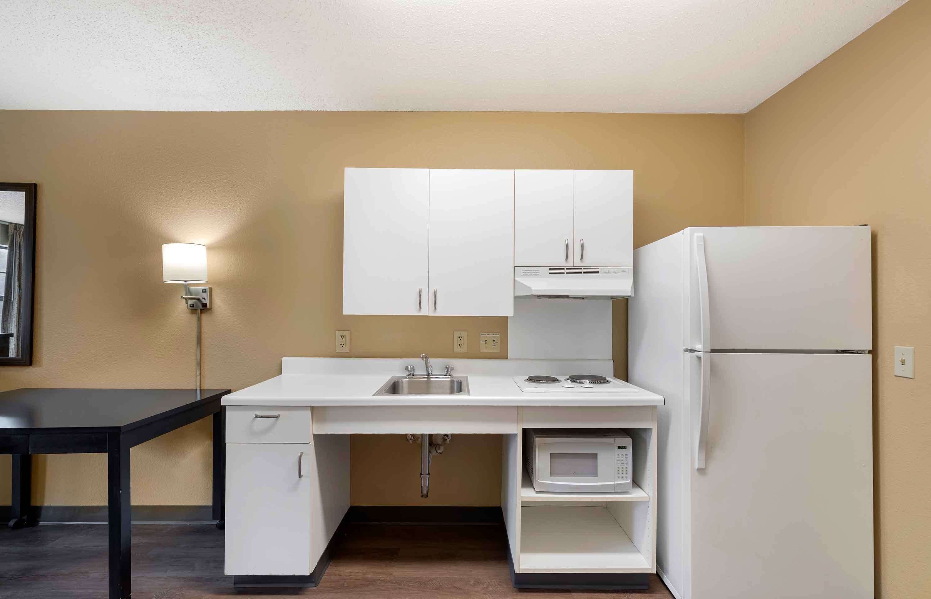 Una kitchenette compacta con armarios blancos, un microondas, un refrigerador, un lavamanos y una pequeña mesa.
