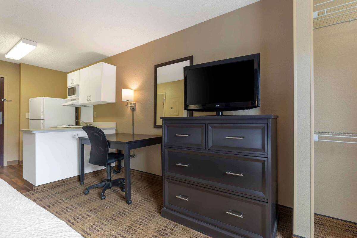 A hotel room with a kitchenette, a flat-screen TV, a desk with a chair, and a dresser.