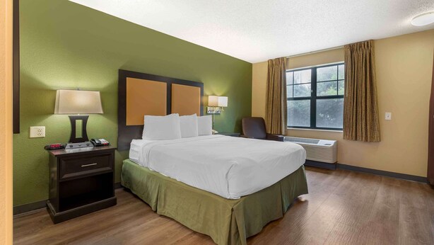 A hotel room with a bed, bedside lamps, a nightstand, a window with curtains, and a thermostat. - Extended Stay America Select Suites - Austin - Northwest - Lakeline Mall (Austin)
