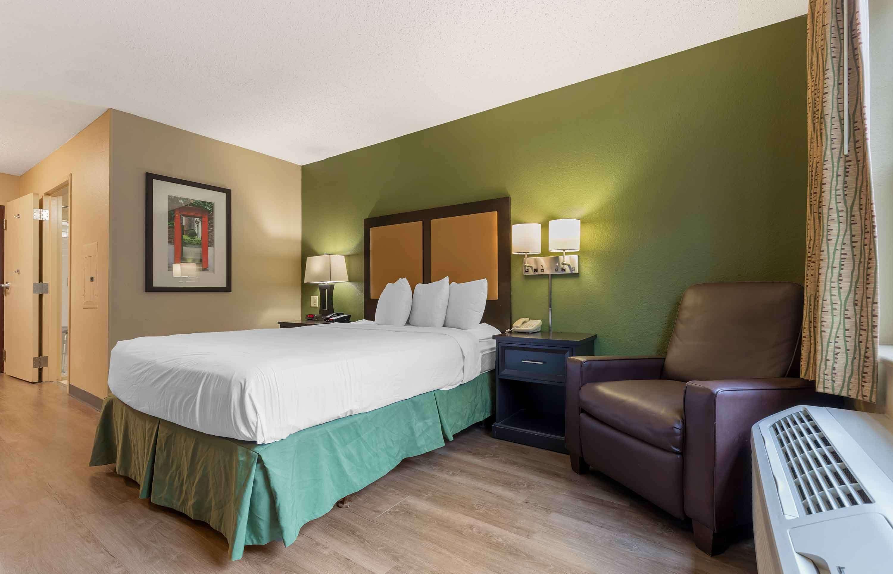 A hotel room with a bed, bedside table, lamp, chair, and a wall-mounted phone.