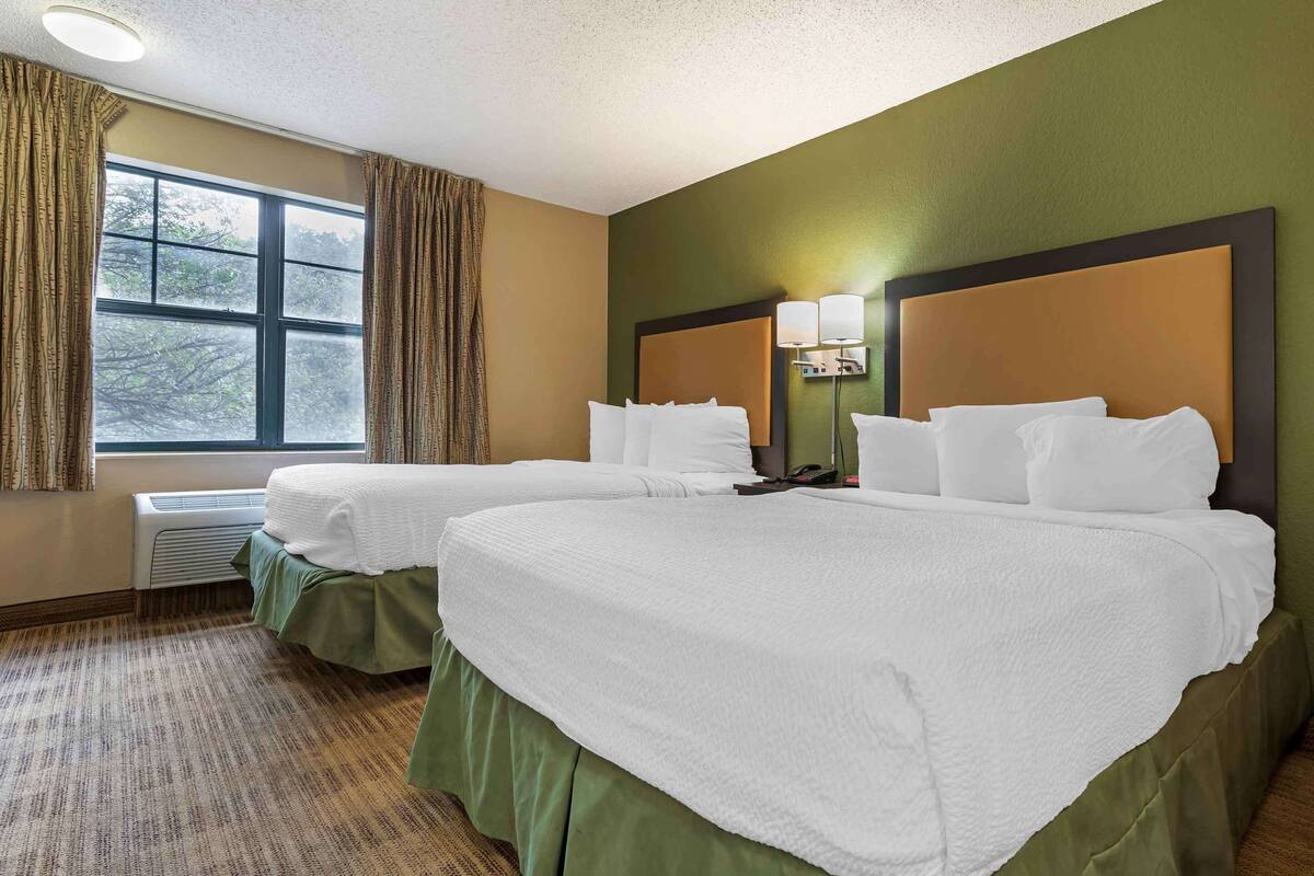 A hotel room with two beds, a large window with curtains, a wall-mounted lamp, and a radiator.
