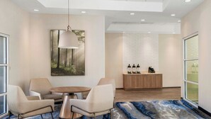 Un hall d’hôtel moderne avec une table ronde, deux chaises, un luminaire suspendu, un tableau, un comptoir d’accueil et une grande fenêtre.