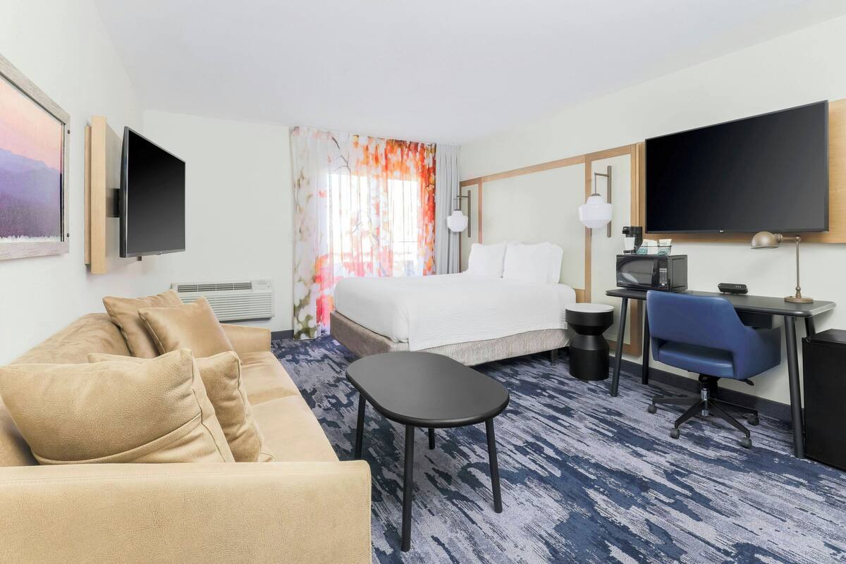 Habitación de hotel con cama, sofá, escritorio con silla, televisor y ventana con cortinas.