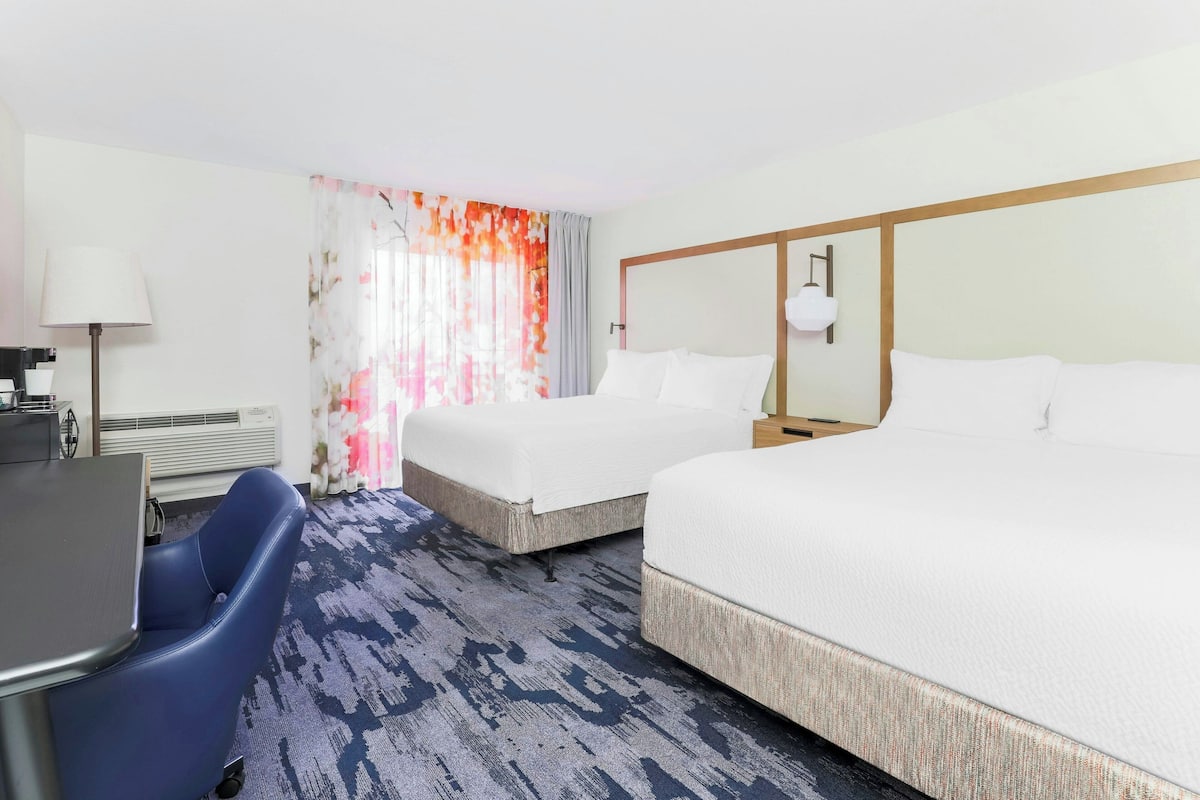 Ein Hotelzimmer mit zwei Betten, einem Schreibtisch, einem Stuhl und einem großen Fenster mit Vorhängen.