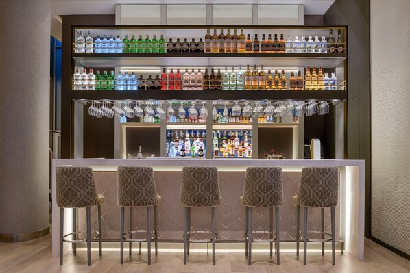 Una zona de bar con múltiples estantes llenos de botellas de licor y vasos, y cuatro taburetes de bar.