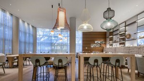 A modern dining area with wooden tables and bar stools, large windows with sheer curtains, and pendant lighting. - AC Hotel Som by Marriott (L'Hospitalet de Llobregat)