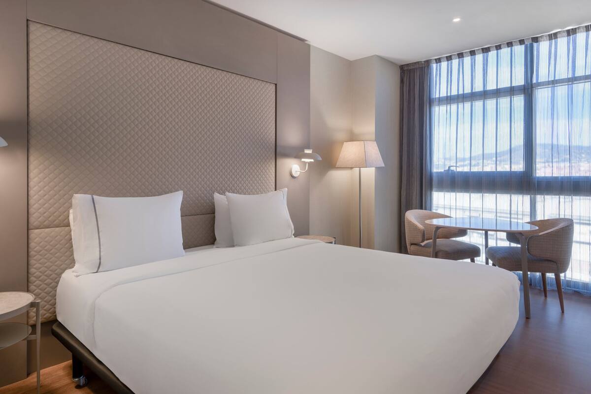 A hotel room with a large bed, a small table with chairs, a lamp, and a view through large windows.