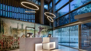 A modern hotel reception area with a large window, a mural of a cityscape, and contemporary lighting fixtures. - AC Hotel Som by Marriott (L'Hospitalet de Llobregat)