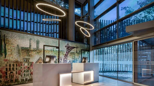 A modern hotel reception area with a large window, a mural of a cityscape, and contemporary lighting fixtures. - AC Hotel Som by Marriott (L'Hospitalet de Llobregat)