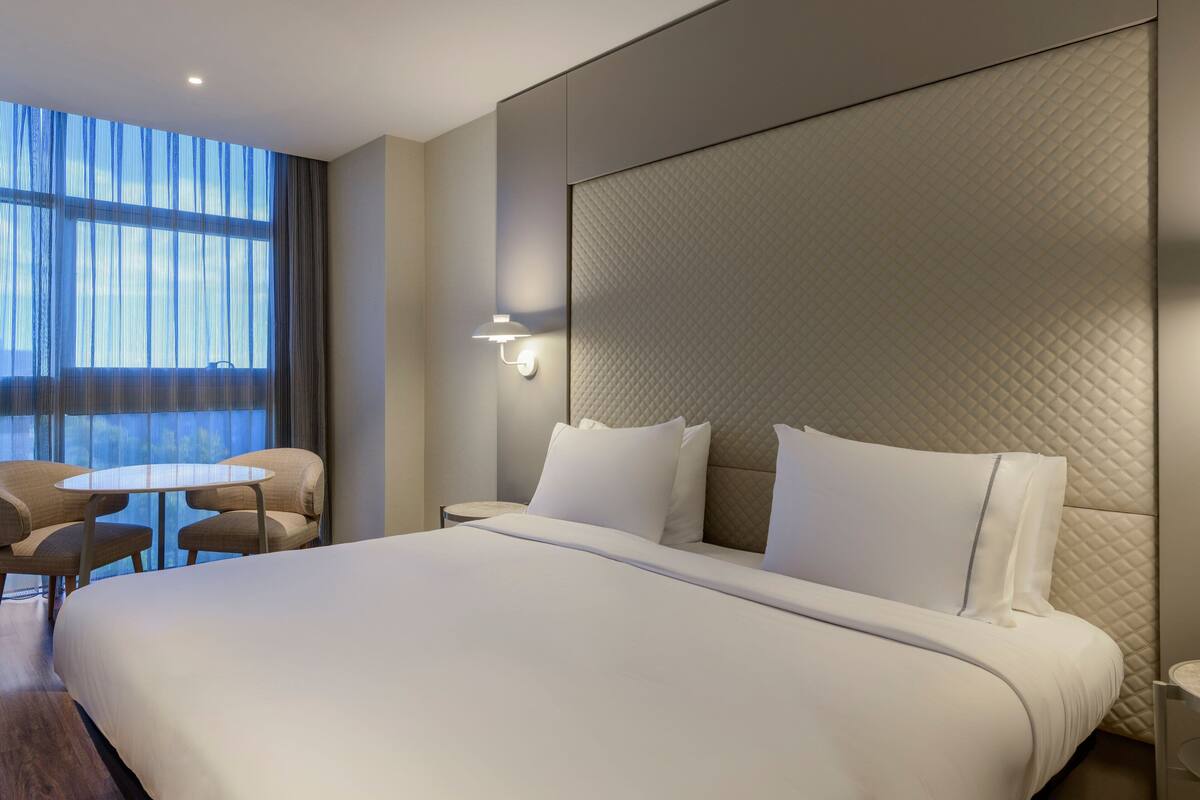 A modern hotel room with a large bed, a small round table, and a view of the outdoors through sheer curtains.