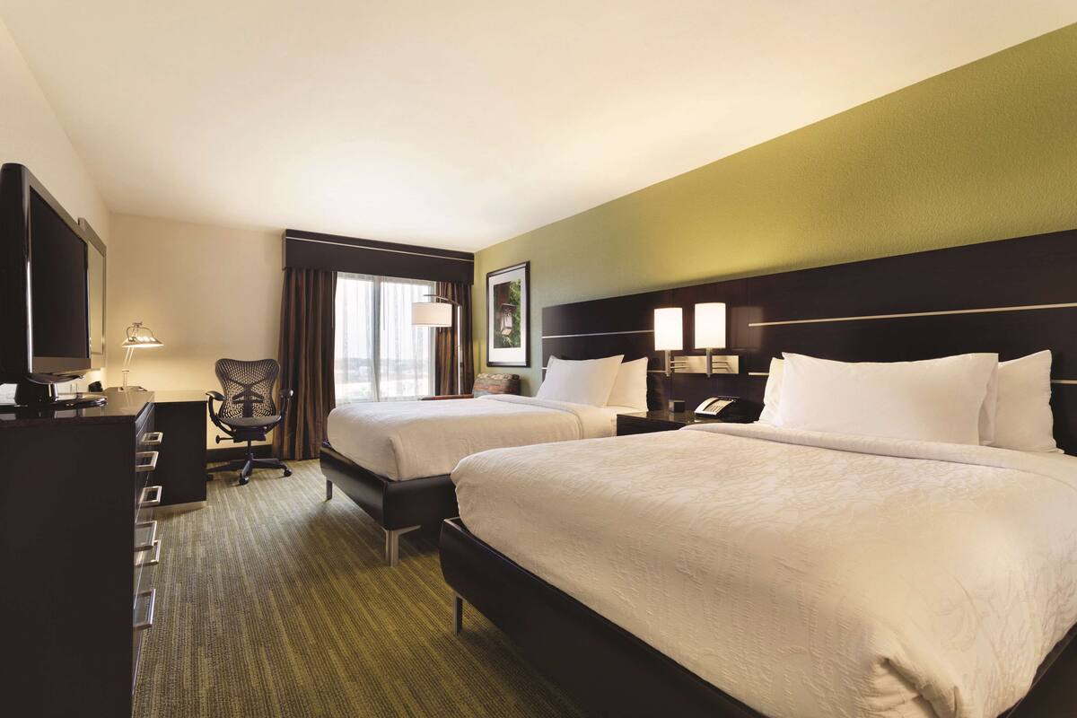 A hotel room with two beds, a desk, a chair, a TV, and a window with curtains.