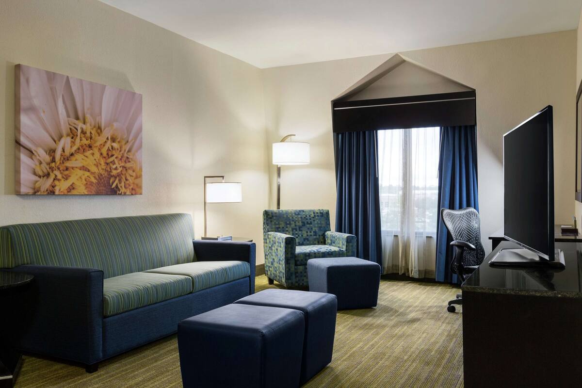 A hotel room with a sofa, two ottomans, a patterned armchair, a desk with a computer, a flat-screen TV, a lamp, and a large window with curtains.