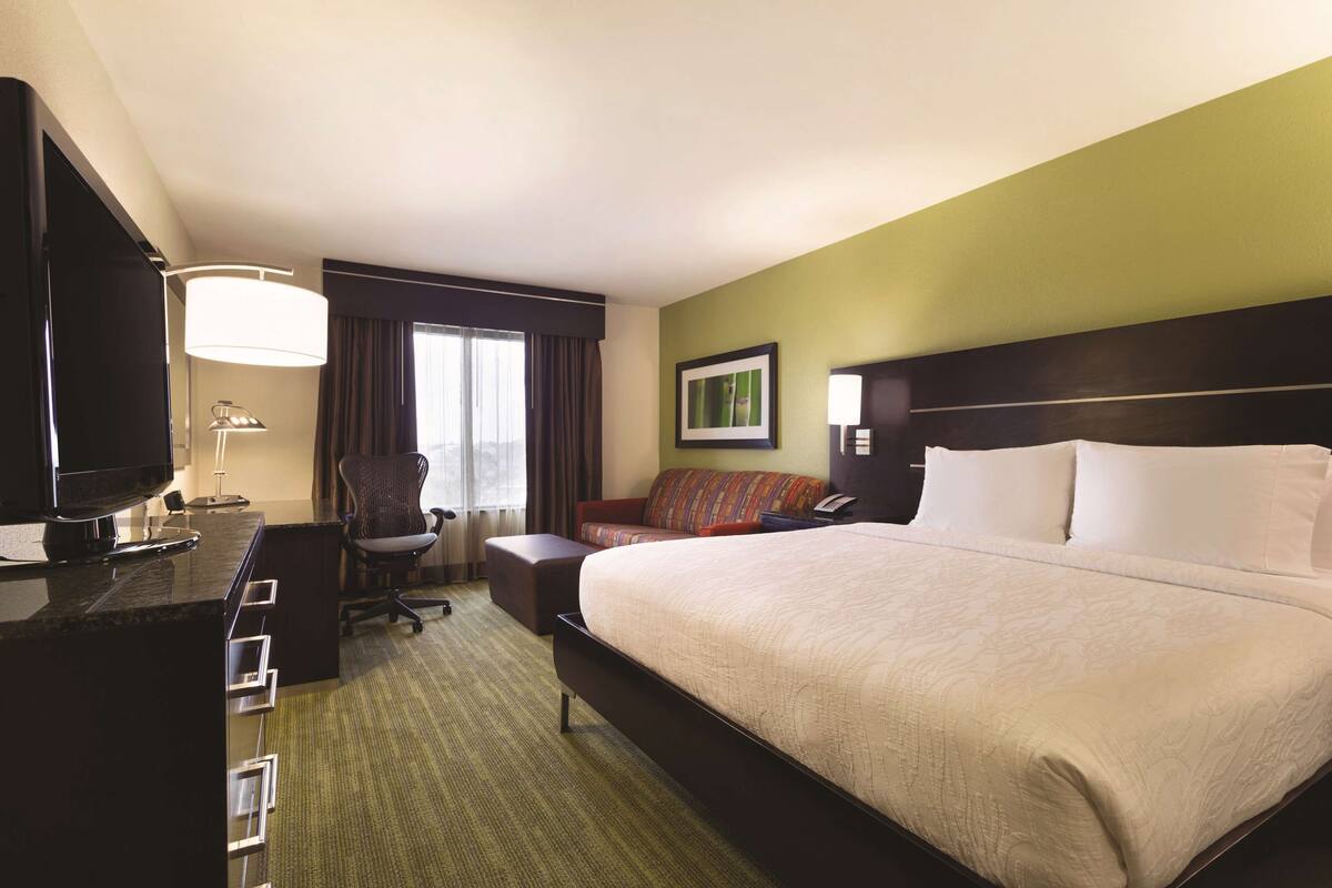 A hotel room with a large bed, a desk with a flat-screen TV, a chair, a sofa, and a window with curtains.
