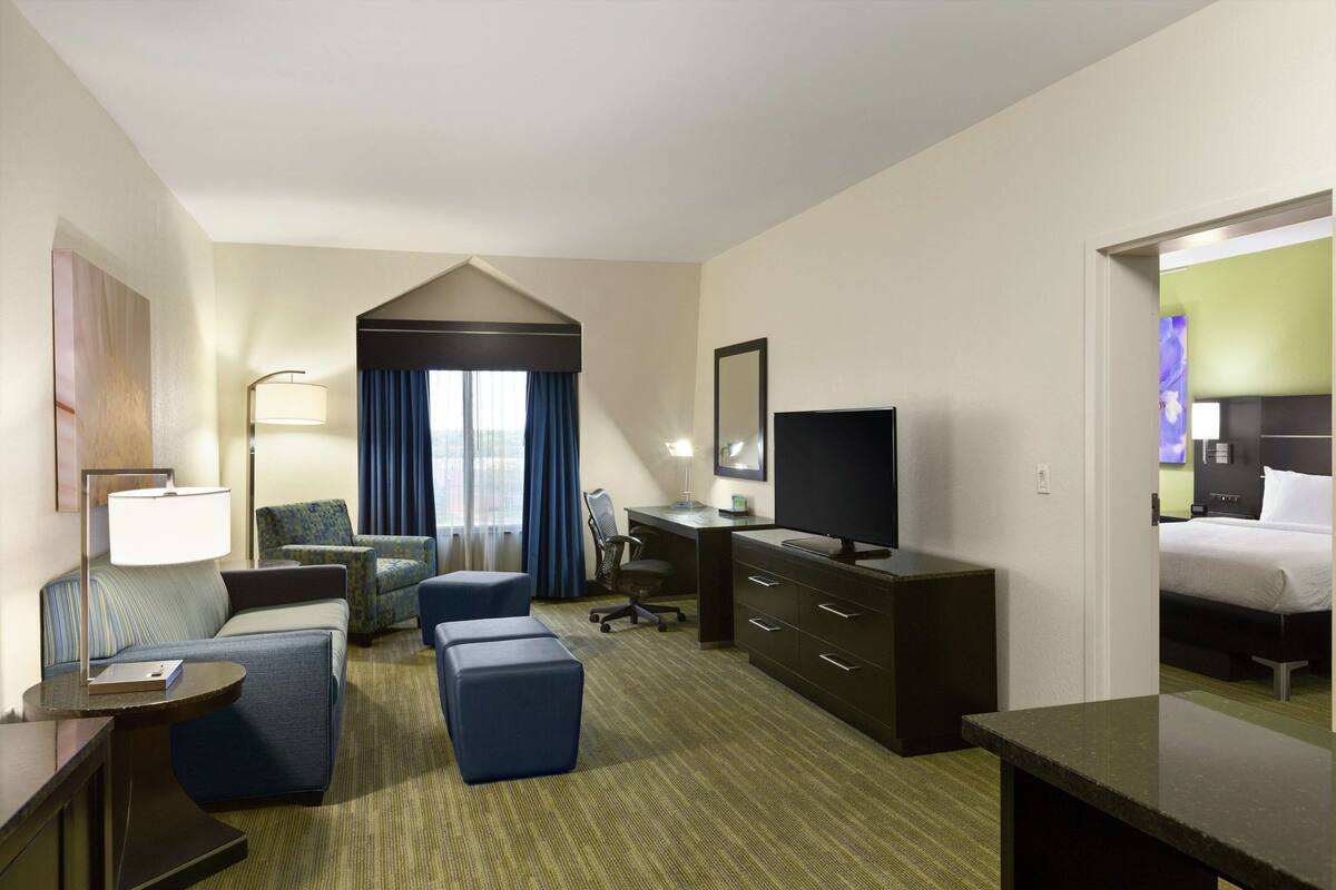 A hotel room with a sofa, armchair, ottoman, desk, TV, and a bed.