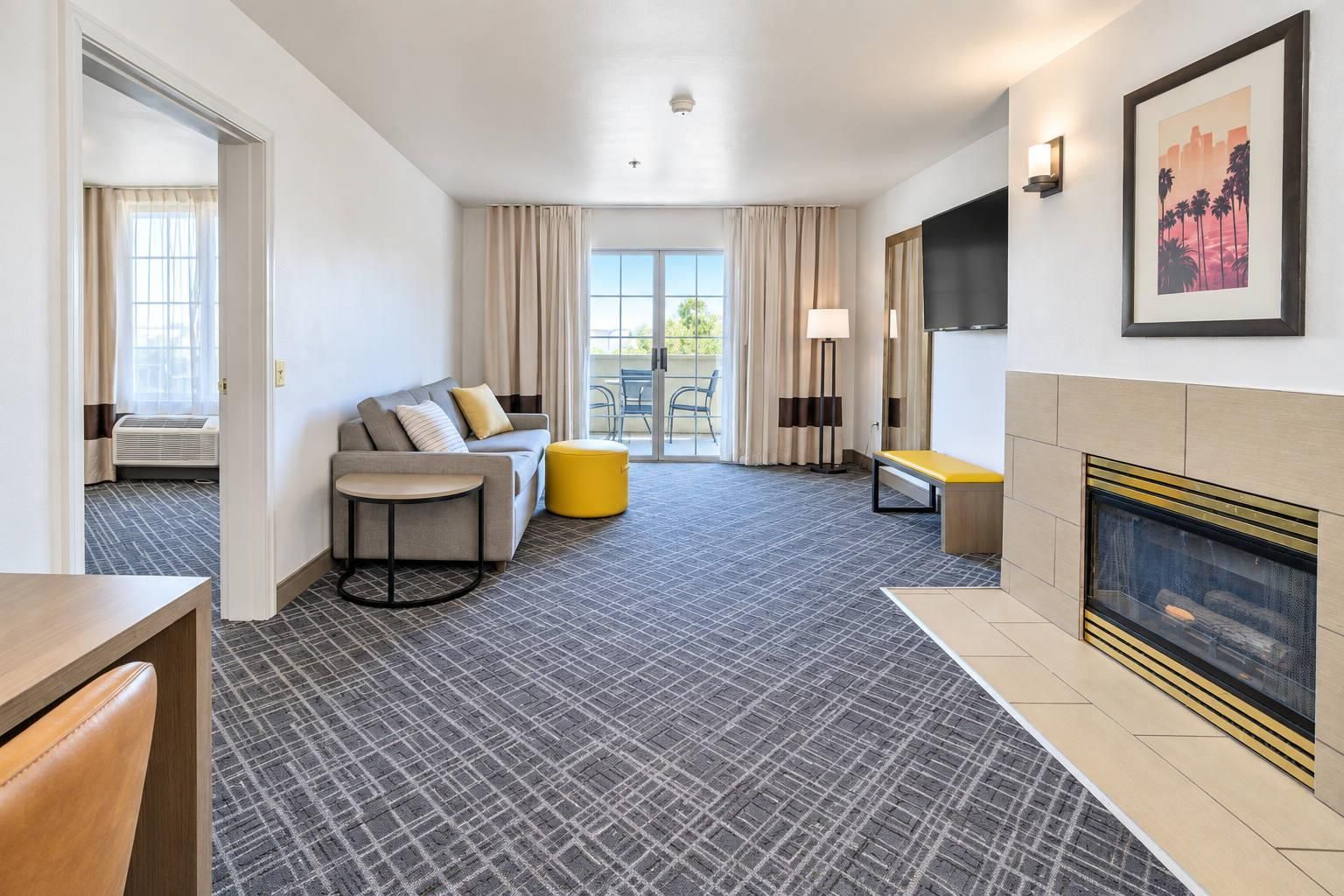A hotel room with a sofa, a yellow ottoman, a flat-screen TV, a fireplace, and a large window with curtains.