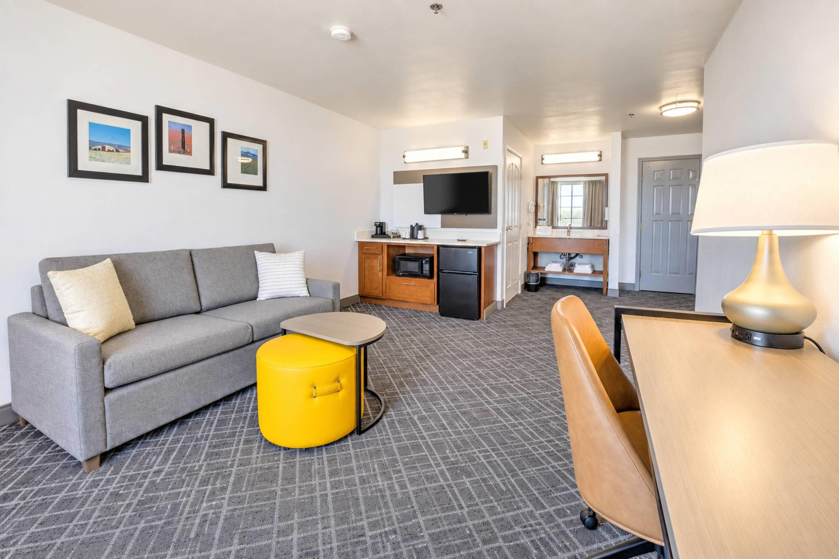 Habitación de hotel moderna con sofá gris, un otoman amarillo, una mesa de madera, un televisor y un baño visible al fondo.
