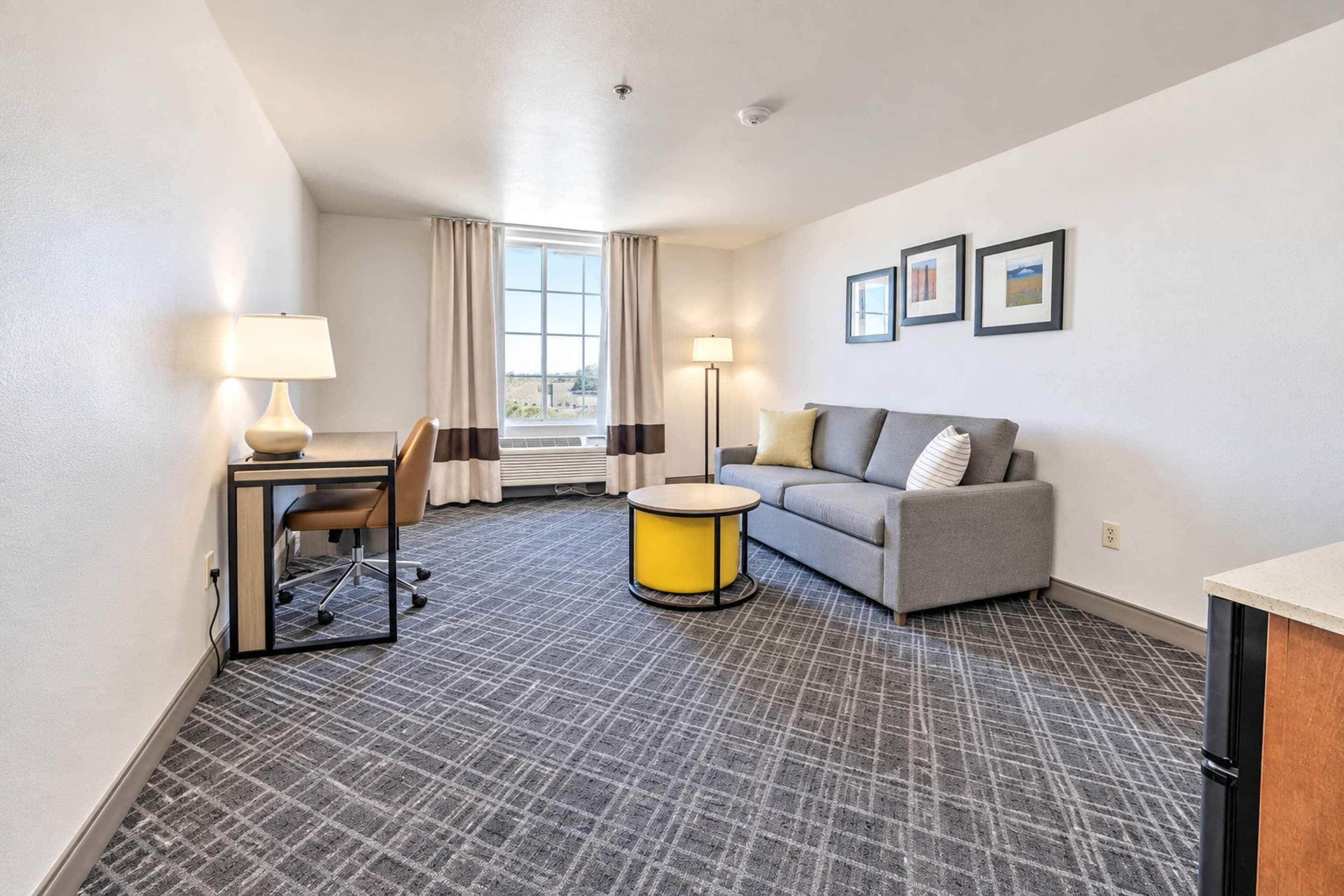 A hotel room with a gray patterned carpet, a gray sofa with a pillow, a small yellow table, a desk with a chair, a lamp, and a window with curtains.