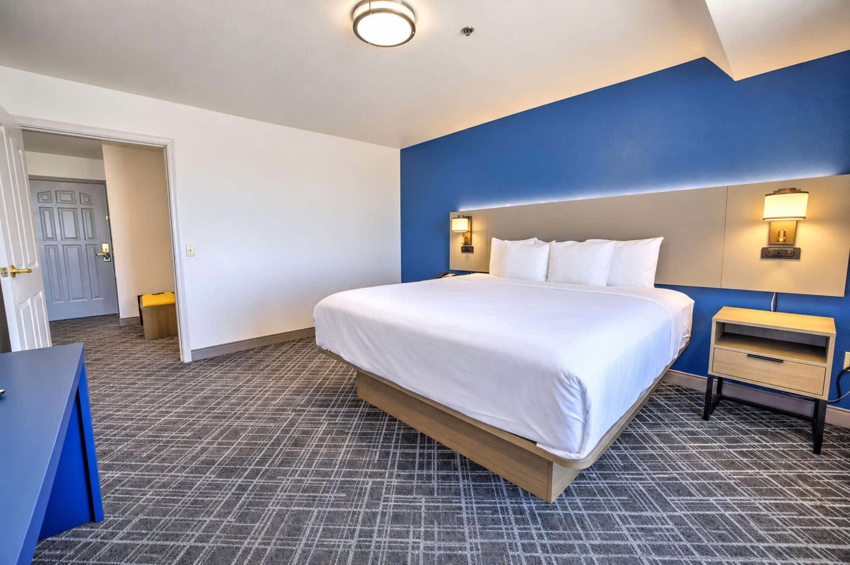 A hotel room with a large bed, a bedside table, a nightstand, and a wall-mounted lamp.