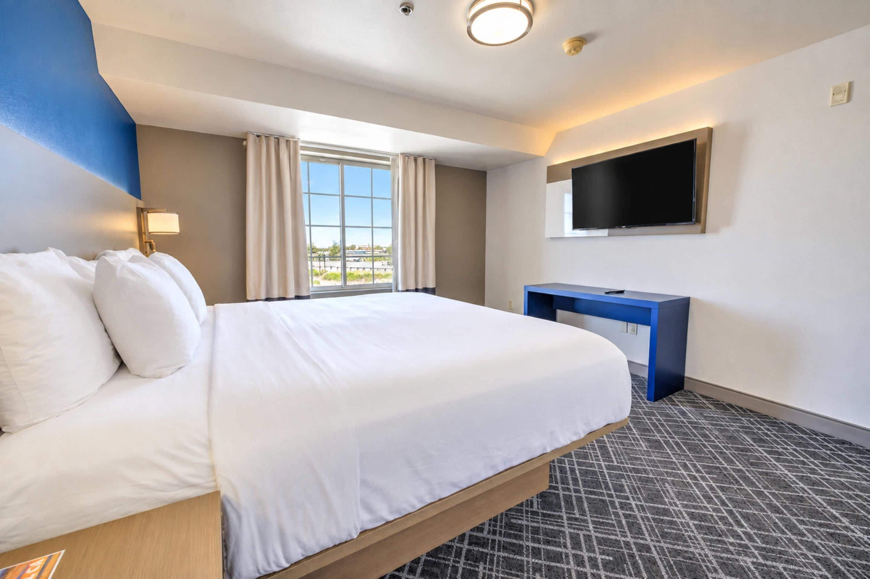 A hotel room with a large bed, a desk, a flat-screen TV mounted on the wall, and a window with curtains.
