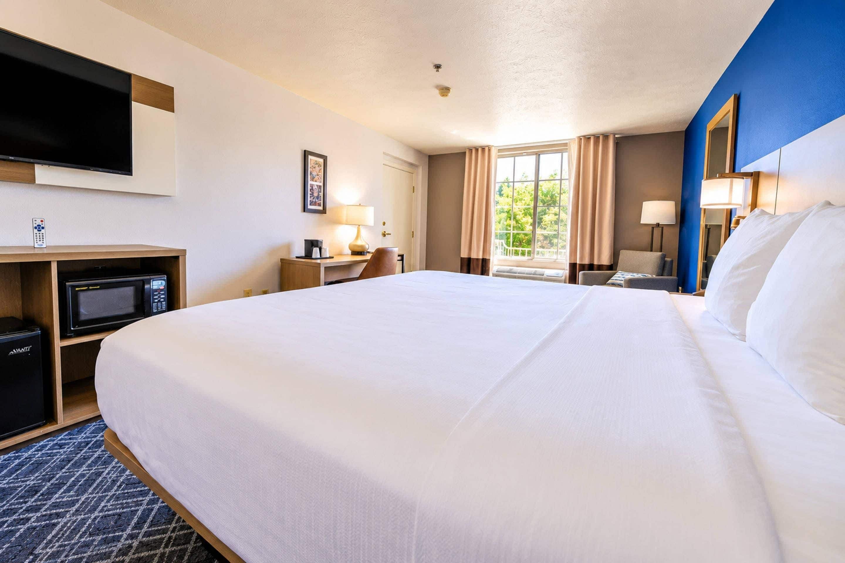 A hotel room with a large bed, a desk with a computer, a TV, and a window with curtains.