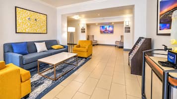A hotel lobby with a blue sofa, yellow armchairs, a coffee table, a reception desk, and a TV screen.