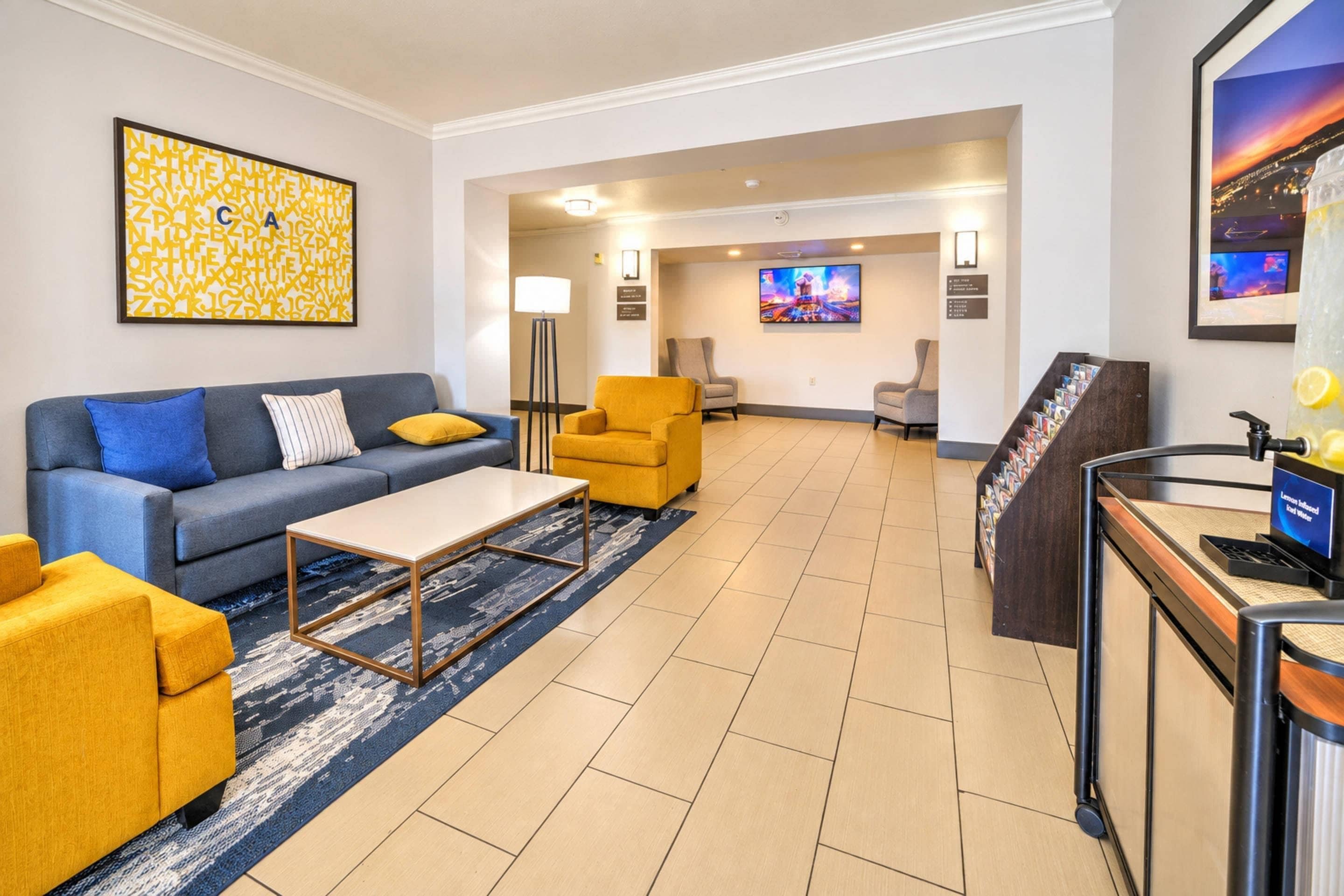 A hotel lobby with a blue sofa, yellow armchairs, a coffee table, a reception desk, and a TV screen.