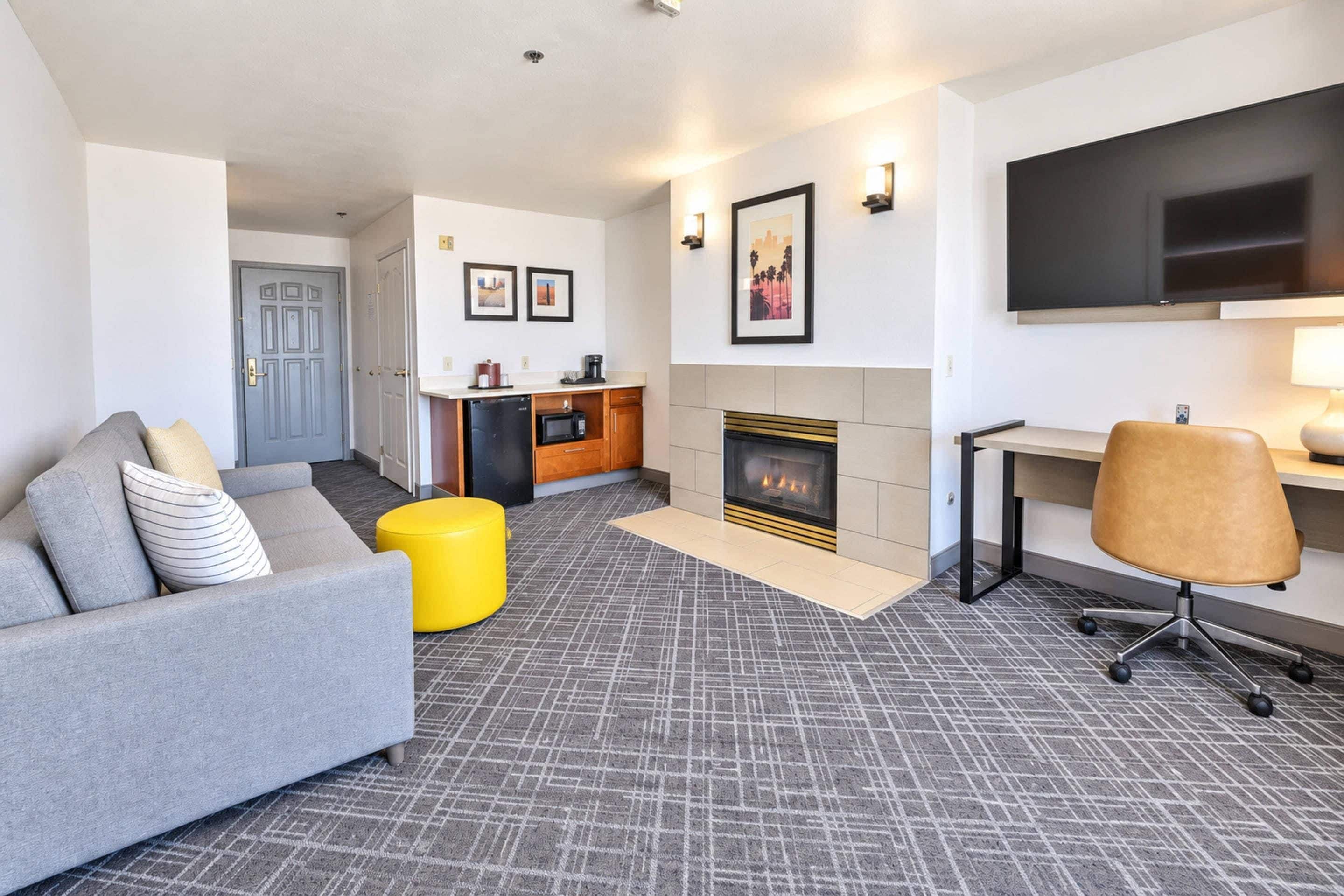 A modern living room with a grey sofa, a yellow ottoman, a fireplace, a flat-screen TV, and a kitchen area with a microwave and coffee maker.
