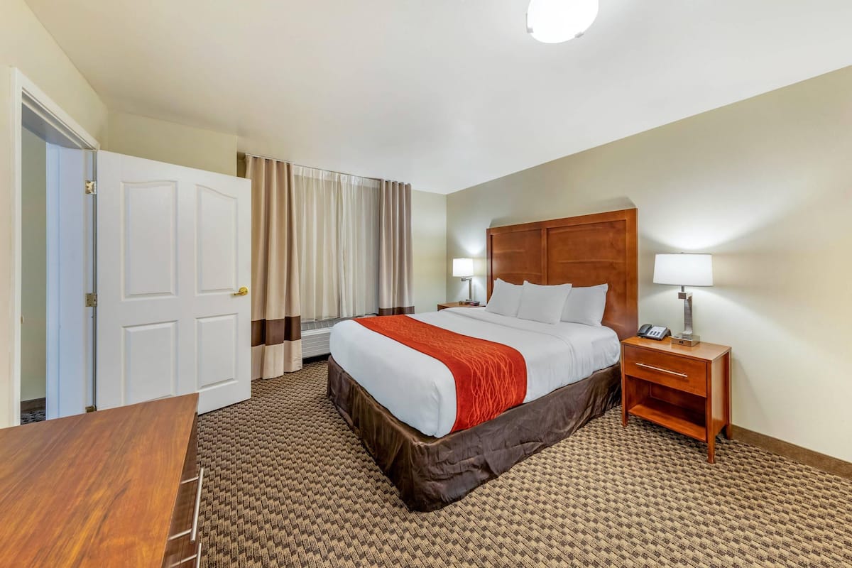 A hotel room with a large bed, a wooden headboard, a bedside table with a lamp, a desk, and a patterned carpet.