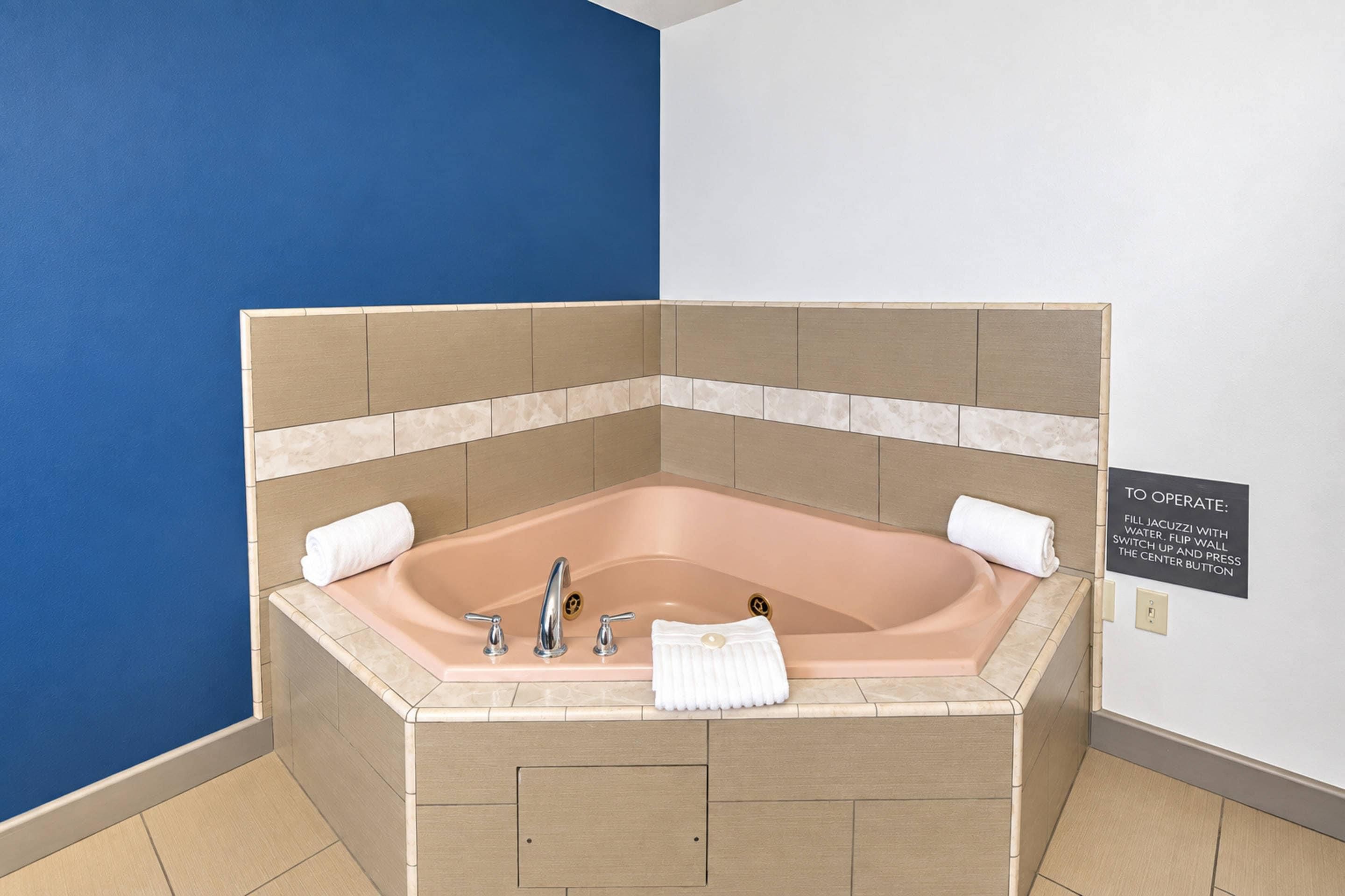 Una bañera en esquina con azulejos color beige, dos llaves y una toalla al lado.