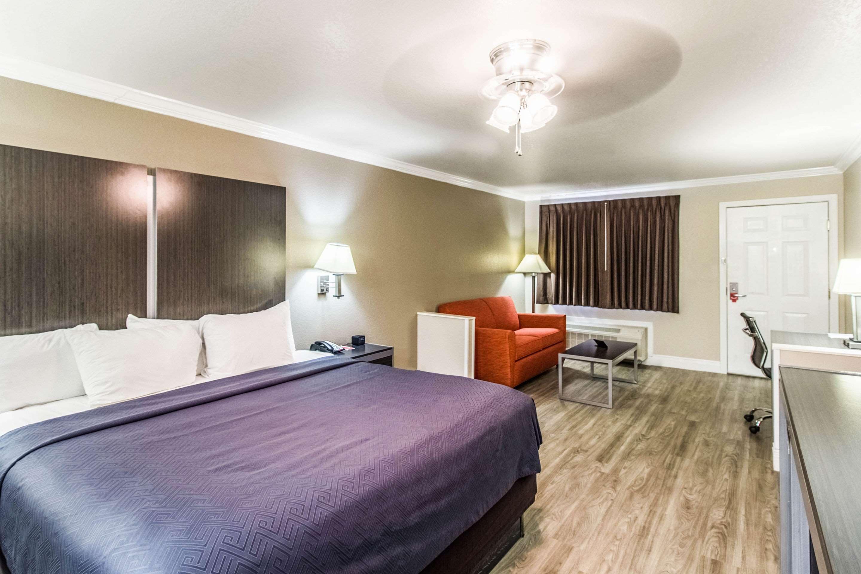 A hotel room with a large bed, a desk, a chair, a desk lamp, a ceiling fan, and a window with curtains.