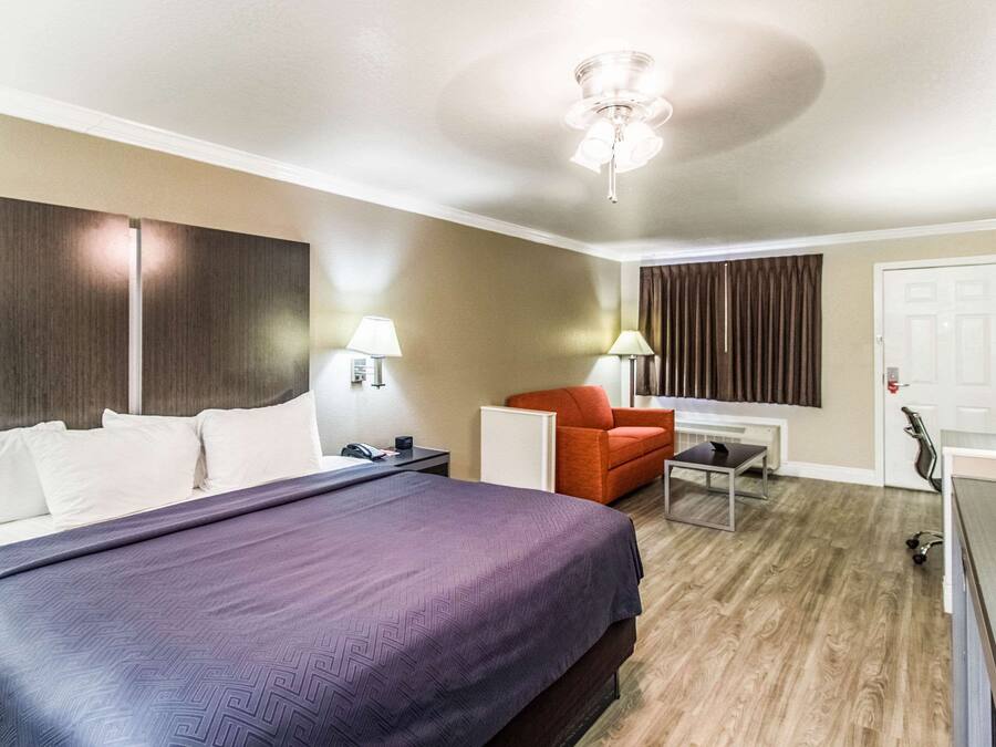 A hotel room with a large bed, a desk, a chair, a desk lamp, a ceiling fan, and a window with curtains.