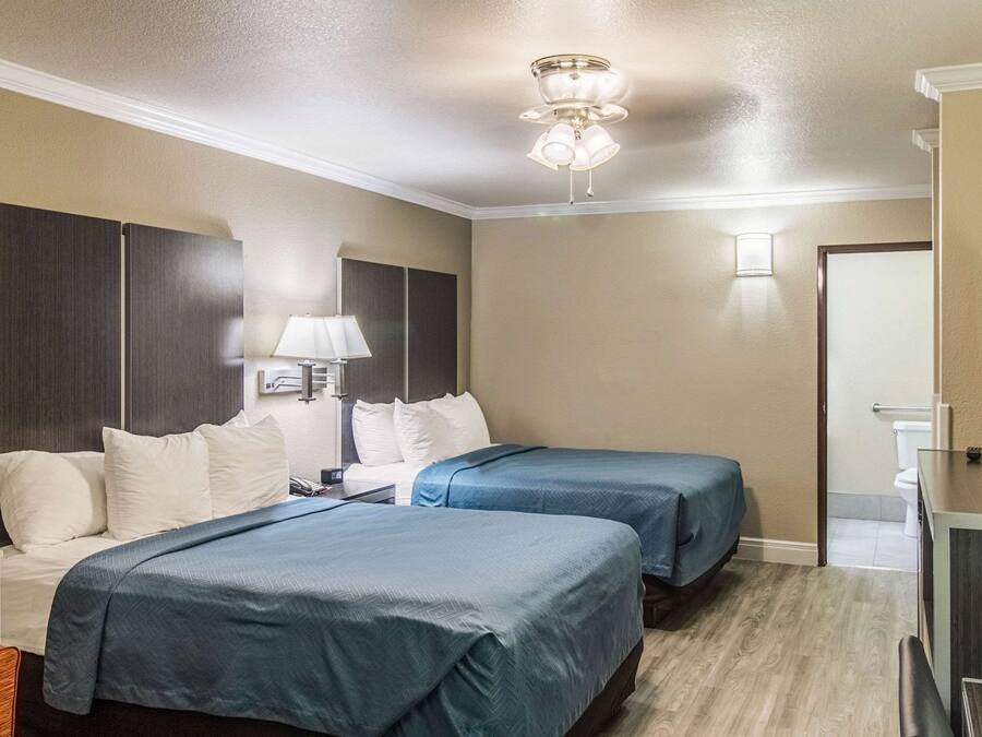 A hotel room with two beds, a desk, a chair, a TV, and a ceiling fan.