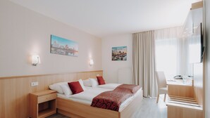 A hotel room with a bed, bedside tables, a desk, a chair, a TV, and two cityscape paintings on the wall. - Hotel Am Wald (Monheim)