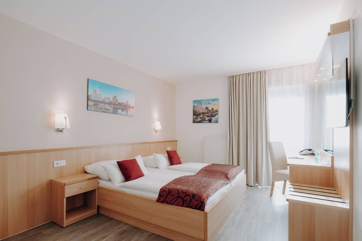 Quarto de hotel com cama, mesas de cabeceira, escrivaninha, cadeira, televisão e duas pinturas de paisagens urbanas na parede.