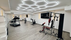 A modern gym with hexagonal ceiling lights, exercise equipment, and a flat-screen TV. - Hotel Am Wald (Monheim)